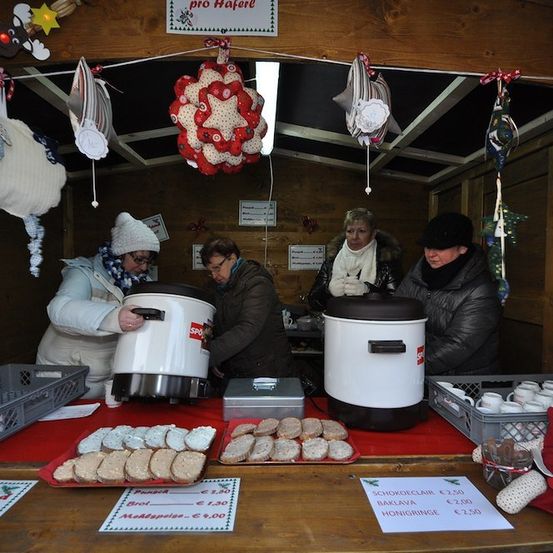 Eine Gruppe von Frauen bereitet in einem kleinen Holzladen Essen zu. Sie stehen um einen Tisch mit Essensartikeln und einem Schild. Der Laden hat eine Holzdecke und Wände. Es hängen Dekorationen und ein Schild mit der Aufschrift 'pro Hafer'. Auf dem Tisch befinden sich auch Tassen und ein Korb.