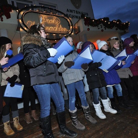 Eine Gruppe von Menschen in Winterkleidung steht vor einem Gebäude, hält blaue Ordner und singt. Das Gebäude hat ein Schild mit der Aufschrift 'Broke nacht'.