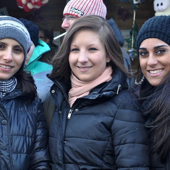 Drei Frauen lächeln und posieren für ein Foto, zwei tragen schwarze Jacken und Mützen, eine trägt einen Schal. Ein Mann, der eine Zigarette raucht, ist im Hintergrund.