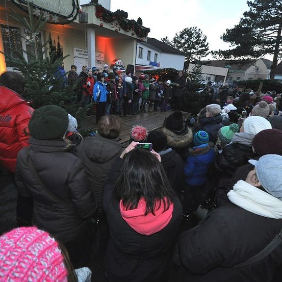 Eine Menschenmenge, darunter Kinder, versammelt sich im Freien in Winterkleidung für eine Veranstaltung vor einem Gebäude mit Weihnachtsdekoration.