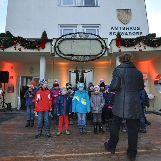 Eine Gruppe von Kindern in Winterkleidung steht auf einer Holzplattform vor einem Gebäude mit festlichen Dekorationen. Eine Person in einem schwarzen Mantel führt sie an.