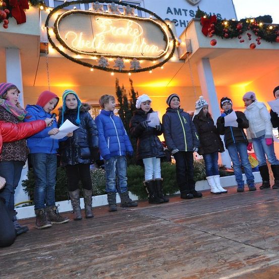 Eine Gruppe von Kindern in Winterkleidung singt Weihnachtslieder auf einer Bühne mit einem Schild, das 'Weihnachtszauber' besagt.