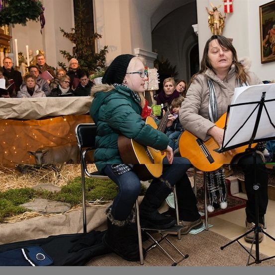 Zwei Frauen spielen Gitarren in einer Kirche, mit einer Krippe im Hintergrund und einem zuschauenden Publikum.