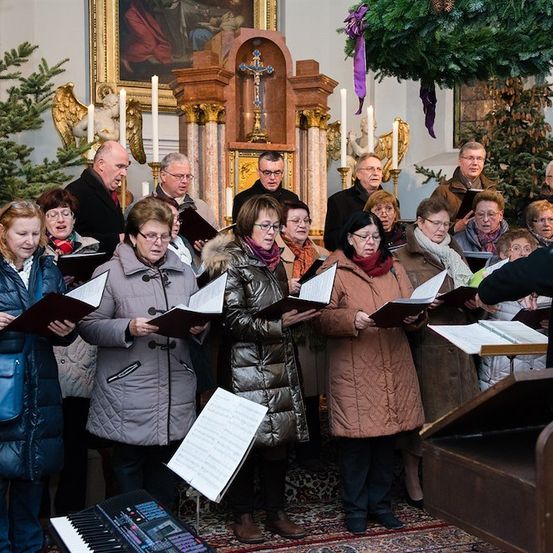 Ein Chor von Menschen in Winterkleidung singt in einer Kirche mit einem Klavier und einem Dirigenten vor ihnen.