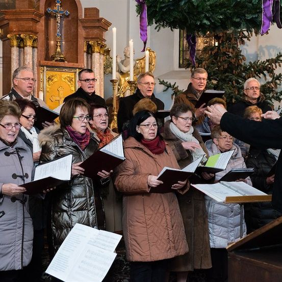 Ein Chor älterer Personen in Wintermänteln singt in einer Kirche, angeführt von einem Dirigenten.