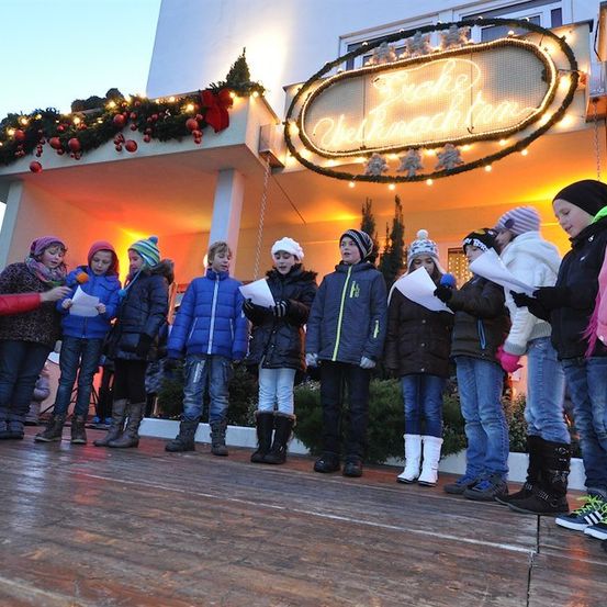 Eine Gruppe von Kindern in Winterkleidung singt Weihnachtslieder vor einem Gebäude mit einem Weihnachtsbaum und einem Schild, das 'Frohe Weihnachten' besagt.