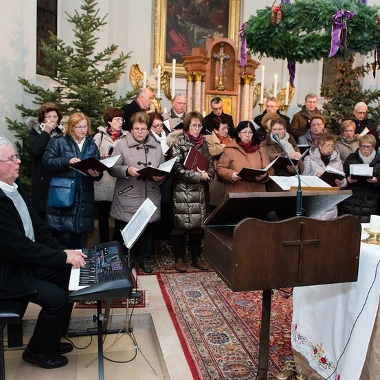 Ein Chor älterer Personen in Winterkleidung singt in einer Kirche, begleitet von einem Mann am Klavier. Die Kirche ist mit Bäumen und Kränzen für Weihnachten geschmückt.