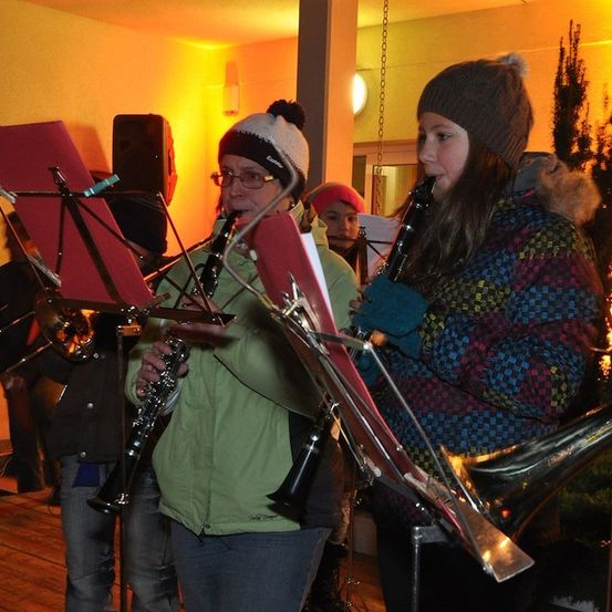Eine Gruppe von Musikern, darunter eine Frau, die Klarinette spielt, und eine andere Frau, die Trompete spielt, treten gemeinsam in einem Raum mit warmer Beleuchtung auf.