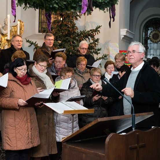 Ein Chor aus Erwachsenen in Winterkleidung steht um ein Podium in einer Kirche, singt, während ein Mann mit Brille und einer schwarzen Jacke dirigiert.