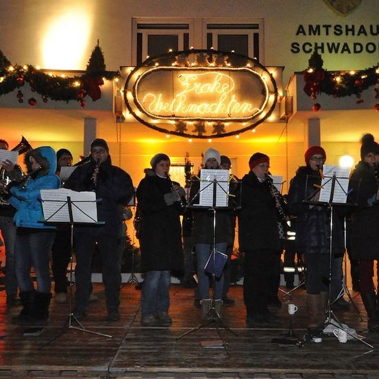 Eine Gruppe von Musikern tritt auf einer Bühne mit festlicher Kulisse auf. Sie spielen verschiedene Instrumente, darunter Trompeten und Klarinetten. Die Bühne ist mit Weihnachtslichtern und einem Schild, das 'Erste Weihnachtsfeier' besagt, geschmückt.