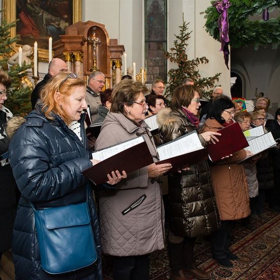 Eine Gruppe von Menschen, möglicherweise ein Chor, singt in einer Kirche. Sie halten Bücher in den Händen und stehen vor einem Weihnachtsbaum und einem Altar.