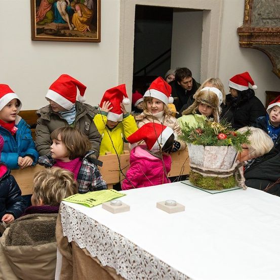 Eine Gruppe von Kindern mit Weihnachtsmützen versammelt sich um einen Tisch mit einer Pflanze und Kerzen. Sie befinden sich in einem Raum mit weißer Wand und einem Gemälde.