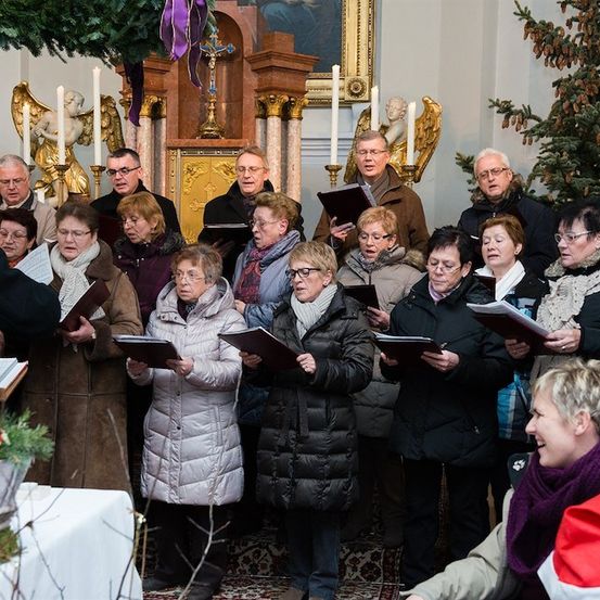 Ein Chor älterer Erwachsener in Winterkleidung singt in einer für Weihnachten geschmückten Kirche. Sie halten Gesangbücher in den Händen und stehen vor einem geschmückten Altar.