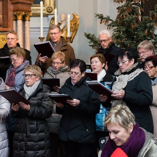Eine Gruppe von Menschen in Winterkleidung versammelt sich in einer Kirche und singt aus Gesangbüchern. Ein Weihnachtsbaum und Kerzen sind im Hintergrund zu sehen.