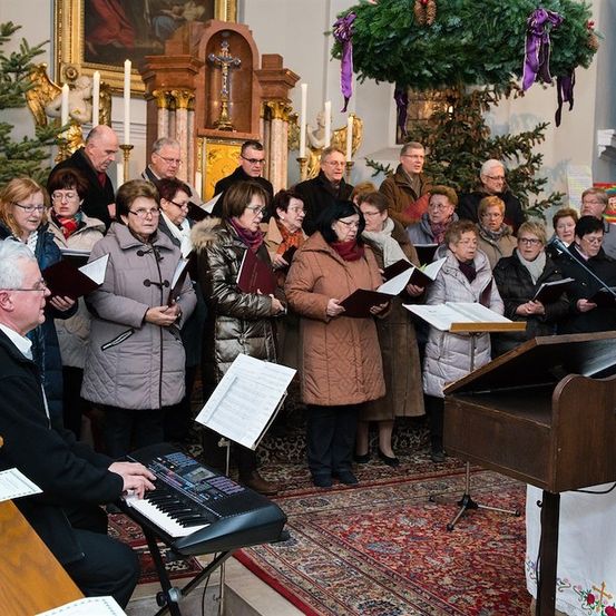 Ein Chor älterer Menschen in Winterkleidung singt in einer Kirche, während ein Mann Klavier spielt.