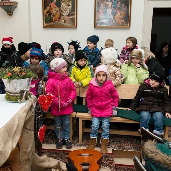 Eine Gruppe von Kindern in Winterkleidung versammelt sich in einem Raum, einige stehend und andere auf Bänken sitzend, mit einer Gitarre und einem Blumenarrangement auf einem Tisch im Vordergrund.