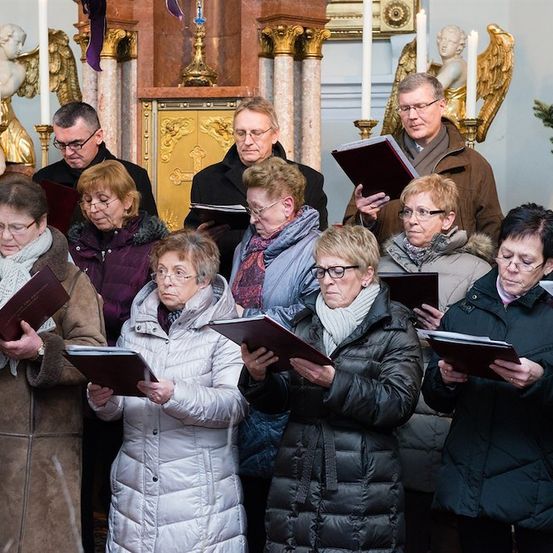 Eine Gruppe von Menschen in Winterkleidung versammelt sich in einer Kirche, hält Gesangbücher in den Händen und singt. Hinter ihnen befinden sich goldene Engelsstatuen und Kerzen auf Ständern.