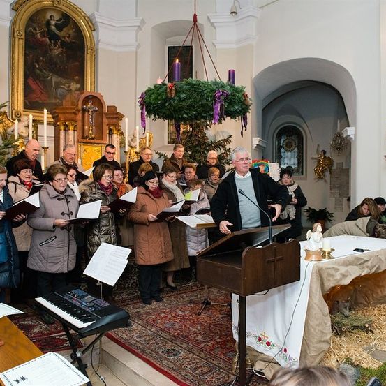 Ein Chor in Winterkleidung singt in einer für Weihnachten geschmückten Kirche. Ein Mann steht an einem Podium mit Mikrofon.