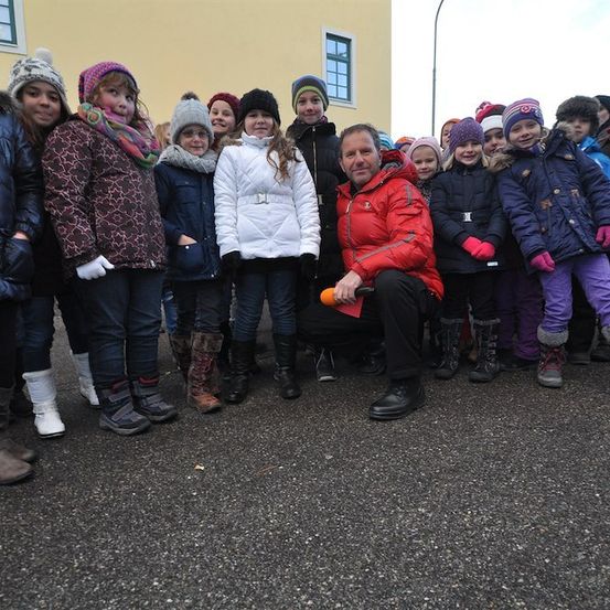 Eine Gruppe von Kindern und ein erwachsener Mann stehen in einer Reihe, lächeln und posieren für ein Foto vor einem Gebäude. Der Mann hält eine Karotte.