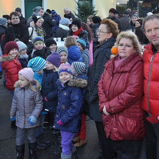 Eine Gruppe von Menschen, einschließlich Erwachsener und Kindern, steht draußen in einer Reihe, möglicherweise bei einer festlichen Veranstaltung. Die Erwachsenen lächeln und einige der Kinder tragen Mützen.