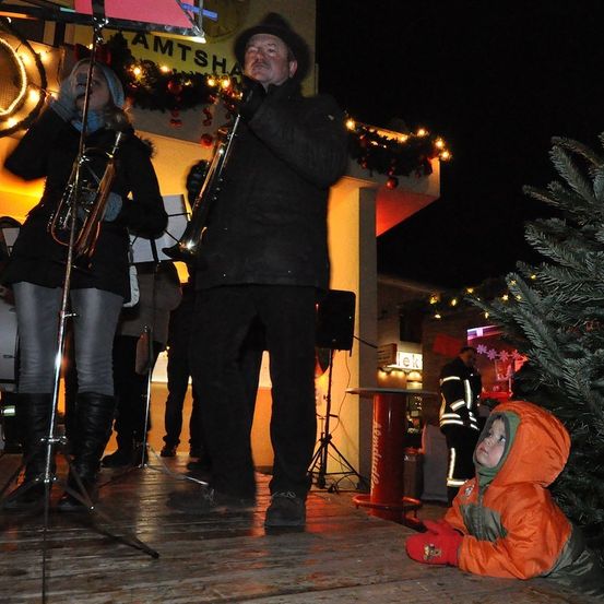 Ein Mann und eine Frau spielen auf einer Bühne bei Nacht Posaunen. Ein Kind in einer orangefarbenen Jacke sitzt in der Nähe auf dem Boden.