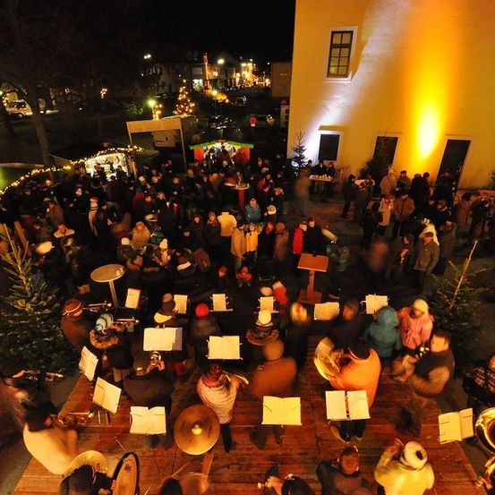 Eine Nachtaufnahme von oben einer festlichen Versammlung mit einer Band, die vor einer Kirche spielt, umgeben von Menschen, Weihnachtsbäumen und festlichen Lichtern.