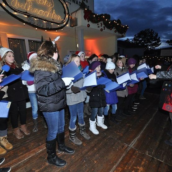 Ein Chor von Kindern in Winterkleidung, die blaue Liederbücher halten, steht auf einer hölzernen Bühne vor einem dekorierten Gebäude bei Nacht.