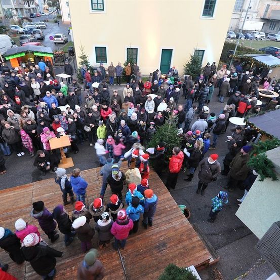 Eine große Menschenmenge, darunter Kinder mit Weihnachtsmützen, versammelt sich auf einer hölzernen Bühne in einer festlichen Umgebung im Freien, mit Gebäuden und geparkten Autos im Hintergrund.