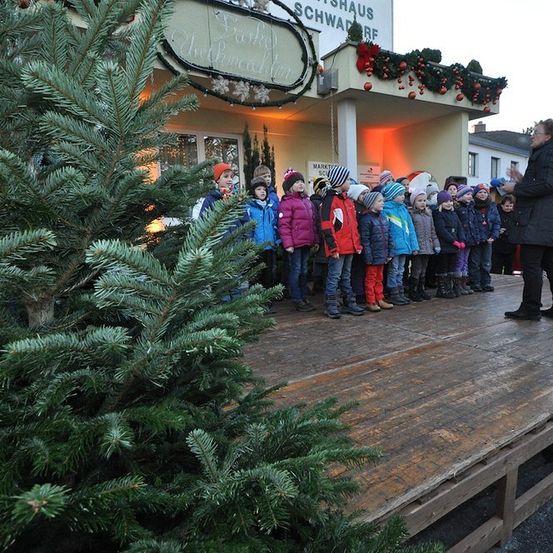 Eine Gruppe von Kindern in Winterkleidung steht auf einer hölzernen Bühne vor einem Weihnachtsbaum, mit einer Frau in der Nähe. Das Gebäude hinter ihnen hat eine Weihnachtsgirlande und Dekorationen.