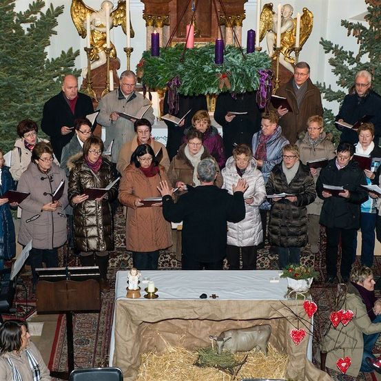 Ein Chor von Menschen in Winterkleidung singt in einer Kirche, mit einer Krippe im Vordergrund.