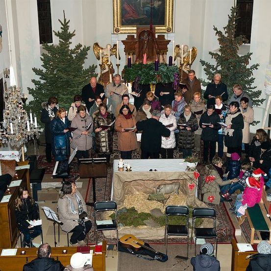 Ein Chor in Winterkleidung singt in einer für Weihnachten geschmückten Kirche, mit einer Krippe und Weihnachtsbäumen im Hintergrund.