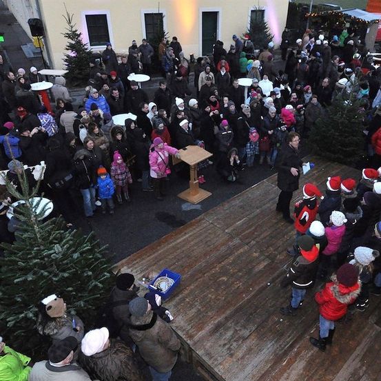 Eine große Menschenmenge, darunter auch Kinder, versammelt sich um eine Holzbühne mit einem Weihnachtsbaum, möglicherweise für eine Feierlichkeit. Einige tragen Mützen und Handschuhe.