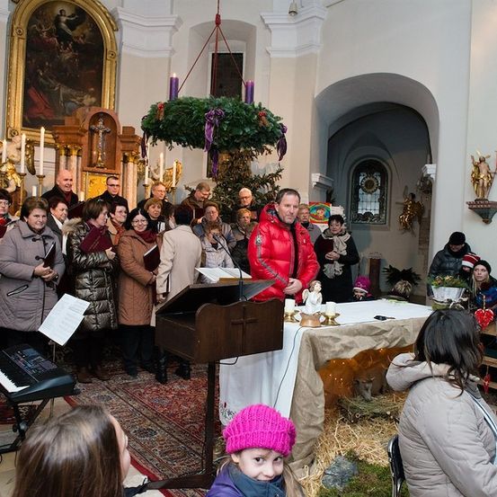 Eine Gruppe von Menschen in einer Kirche, einige stehend, andere sitzend, mit einem Mann in einer roten Jacke an einem Podium, einem Klavier und einer Krippe auf einem Tisch.