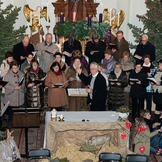 Ein Chor von Menschen in Winterkleidung steht vor einem Kirchenaltar und singt während eines Weihnachtsgottesdienstes. Der Altar ist mit einer Krippe, Kerzen und Weihnachtsdekoration geschmückt.