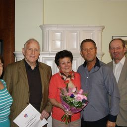 Fünf Menschen stehen eng beieinander und lächeln für ein Foto. Eine Frau in der Mitte hält einen Blumenstrauß. Der Mann auf der linken Seite hält ein Zertifikat. Hinter ihnen steht ein weißer Kamin.