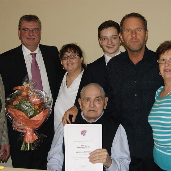 Eine Gruppe von Menschen um einen älteren Mann, der ein Zertifikat in der Hand hält, mit einem Blumenstrauß davor. Sie alle lächeln und scheinen einen besonderen Anlass zu feiern.