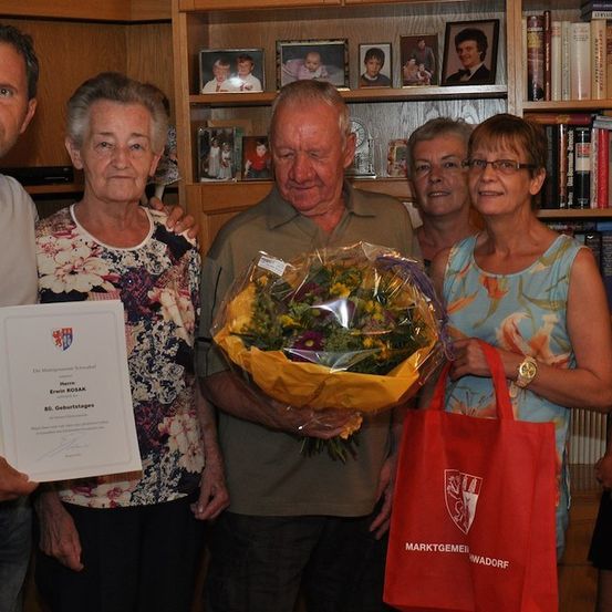 Eine Gruppe von Menschen feiert einen Geburtstag. Ein älterer Mann hält einen Blumenstrauß. Eine Frau hält ein Zertifikat und eine rote Tasche.