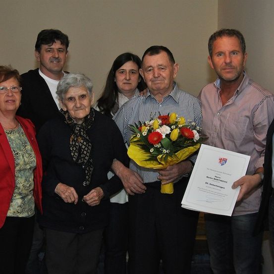 Eine Gruppe von Menschen steht zusammen und lächelt für ein Foto. Eine ältere Frau hält einen Blumenstrauß und ein Mann hält ein Zertifikat.