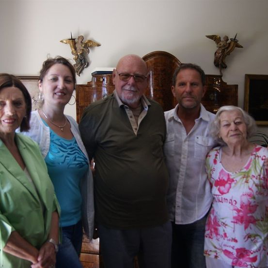Fünf Menschen posieren für ein Foto in einem Raum mit weißen Wänden. Die Frau rechts trägt ein Blumenhemd. Der Mann in der Mitte trägt eine Brille. Hinter ihnen stehen Holzmöbel und Bilderrahmen an der Wand.