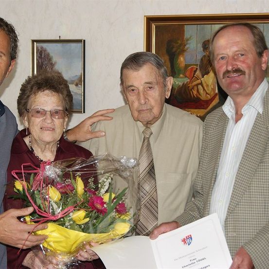 Vier Menschen stehen zusammen, zwei Männer auf beiden Seiten eines älteren Paares, die Frau hält einen Blumenstrauß und der Mann ein Zertifikat.
