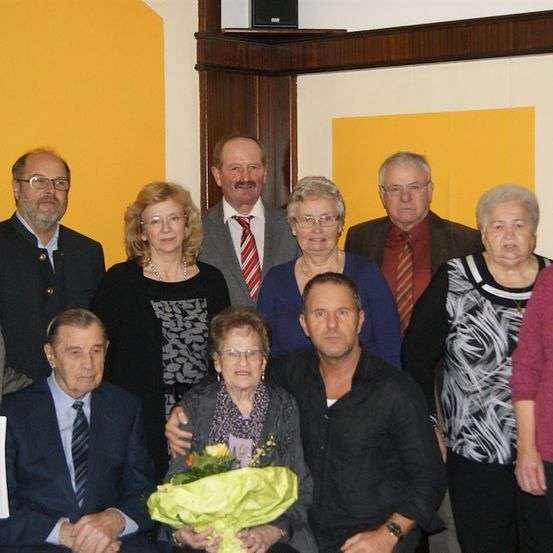 Eine Gruppe von formell gekleideten Menschen posiert für ein Foto. Eine ältere Frau hält einen Blumenstrauß. Ein Mann in einem schwarzen Shirt sitzt mit seinem Arm um die Frau. Ein Zertifikat ist im Vordergrund sichtbar.