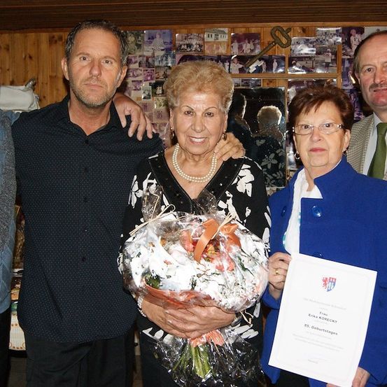 Fünf Menschen stehen zusammen und lächeln für ein Foto. Eine Frau hält einen Blumenstrauß und ein Zertifikat. Der Mann auf der linken Seite hat seinen Arm um ihre Schultern gelegt. Hinter ihnen ist eine Wand mit Bildern und einem Schlüssel.