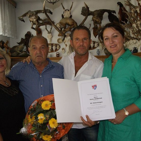 Vier Personen stehen vor einer Wand mit verschiedenen Trophäen und einem Zertifikat, das von einem Mann gehalten wird. Die Frau rechts lächelt und hält einen Blumenstrauß.