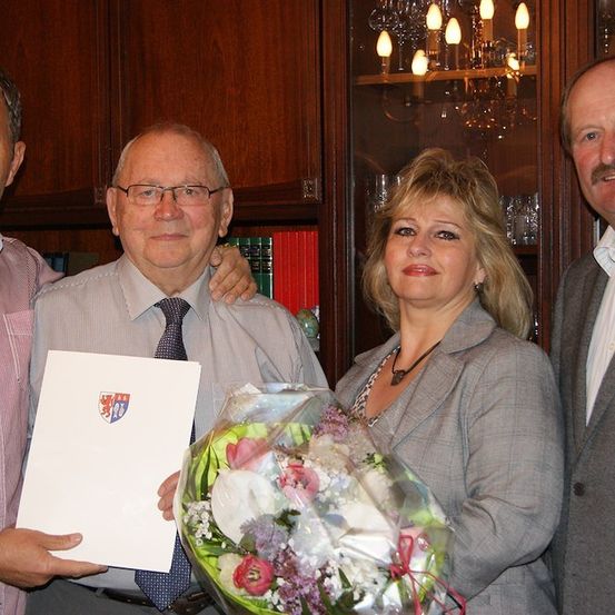 Ein Mann, der ein Zertifikat und einen Blumenstrauß hält, steht mit zwei anderen Erwachsenen in einem Raum mit Bücherregalen und einer Kronleuchter.