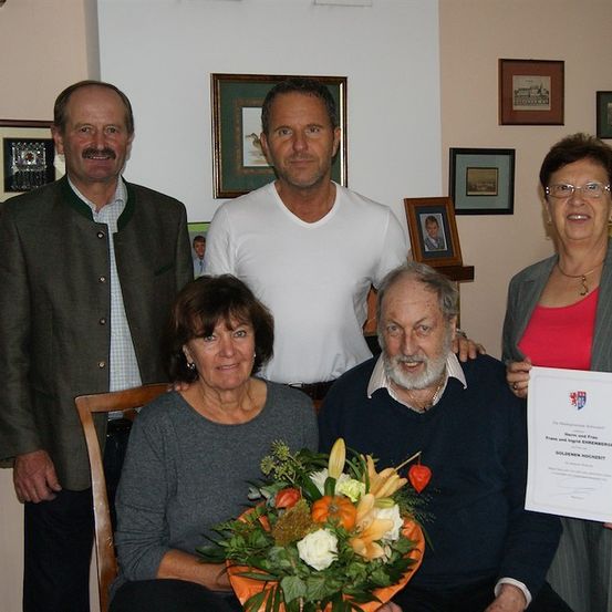 Fünf Personen sind in einem Raum versammelt und lächeln für ein Foto. Ein Mann und eine Frau sitzen auf einem Stuhl mit einem Blumenarrangement vor ihnen. Ein weiterer Mann steht hinter ihnen und zwei Frauen stehen auf beiden Seiten. Eine der Frauen hält ein Zertifikat.