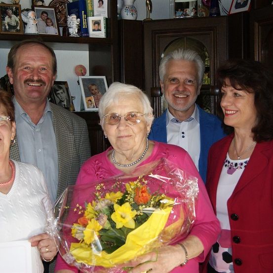 Eine Gruppe von Menschen posiert für ein Foto, wobei eine ältere Frau einen Blumenstrauß hält. Hinter ihnen steht ein Holzregal mit Bilderrahmen und anderen Gegenständen.