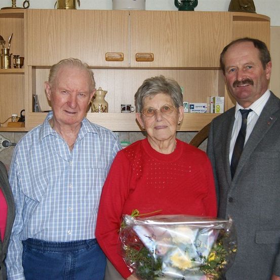 Vier Menschen stehen zusammen in einem Raum und lächeln für ein Foto. Die Frau in der Mitte hält einen Blumenstrauß. Hinter ihnen befinden sich Holzschränke mit verschiedenen Gegenständen auf den Regalen.