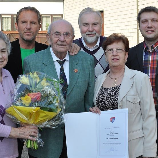Eine Gruppe von Menschen steht für ein Foto zusammen. Ein älterer Mann hält einen Blumenstrauß und ein Zertifikat. Eine Frau auf der linken Seite hält den Strauß. Eine Frau auf der rechten Seite hält ein Zertifikat. Hinter ihnen trägt ein Mann auf der rechten Seite ein kariertes Hemd.