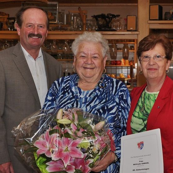 Eine ältere Frau in einem blau-weiß gemusterten Oberteil hält einen Strauß rosa Lilien und ein Zertifikat, flankiert von zwei lächelnden Personen in einem Raum mit Regalen im Hintergrund.
