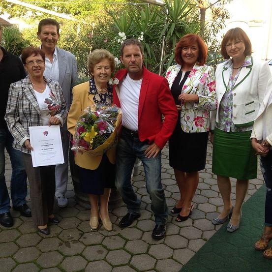 Eine Gruppe von Menschen steht draußen und lächelt für ein Foto. Eine Frau hält einen Blumenstrauß und ein Mann in einem roten Jackett hält ihre Hand. Hinter ihnen hält eine Frau ein Zertifikat.